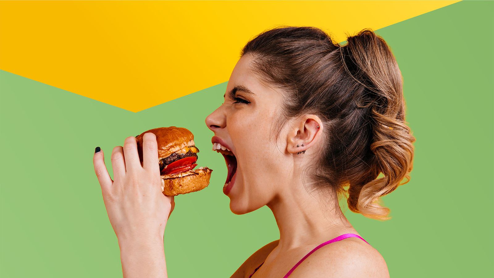 Junge Frau beisst mit Vorfreude in einen saftigen Burger (mit gelbem Designelement oberhalb)
