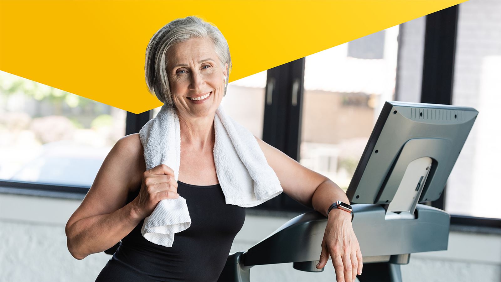 Eine ältere sportliche Frau mit Sportbekleidung und Handtuch steht vor einem Hometrainer in einem Fitnessstudio.