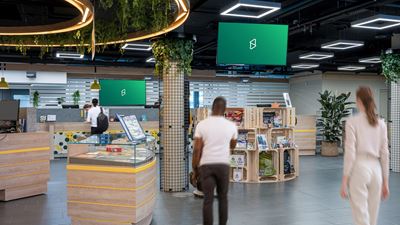 Vue intérieure d’un espace de vente ou de séjour moderne avec plusieurs écrans numériques suspendus au plafond; au premier plan, on voit des personnes en mouvement et différentes surfaces de présentation.