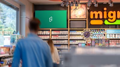 Écran publicitaire numérique dans l’espace de vente d’un kiosque ou d’un convenience shop; au premier plan, on voit des clients flous et des rayons avec des produits.