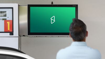 Écran publicitaire numérique dans une station-service, à côté de la pompe; au premier plan, on voit une personne de dos.