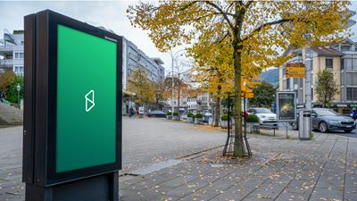 Écran publicitaire numérique dans l’espace public, le long d’une rue ou dans un centre-ville; à l’arrière-plan, on voit des arbres, des bâtiments et des voitures en circulation.