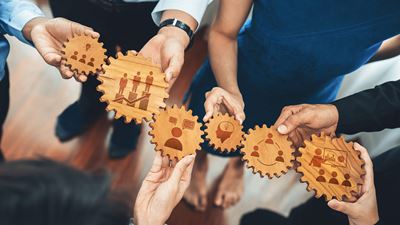 Eine Gruppe von Menschen hält Zahnräder aus Holz mit verschiedenen Icons in den Händen und steht symbolisch für Teamarbeit, Innovation und strategische Planung.